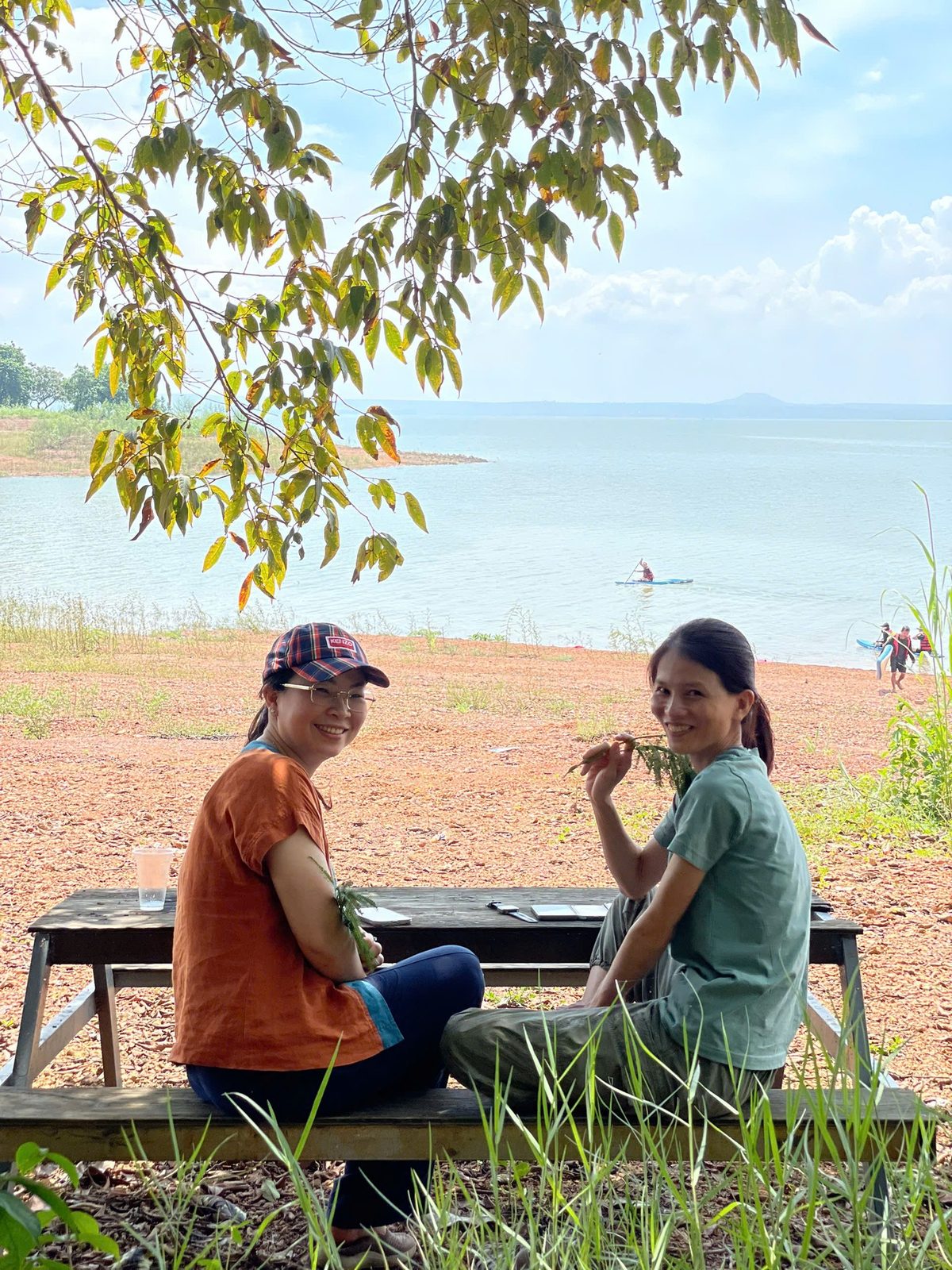 Hai cô gái ngồi bàn picnic ven hồ, người chèo SUP phía sau